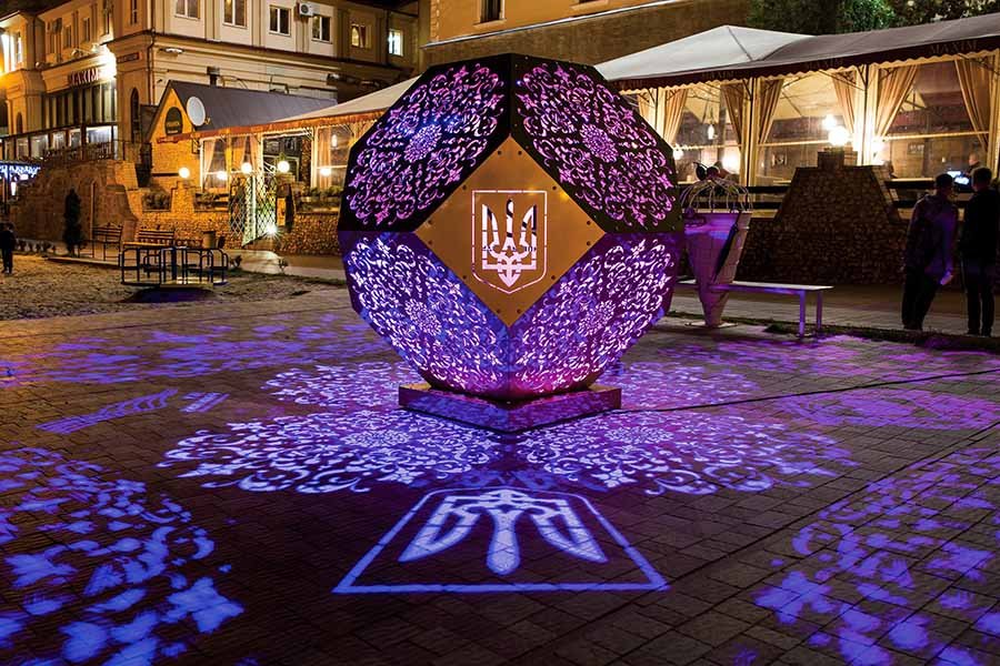 illuminated geometric sculpture projecting floral patterns onto the ground in a public night installation near a café terrace illuminated geometric sculpture projecting floral patterns