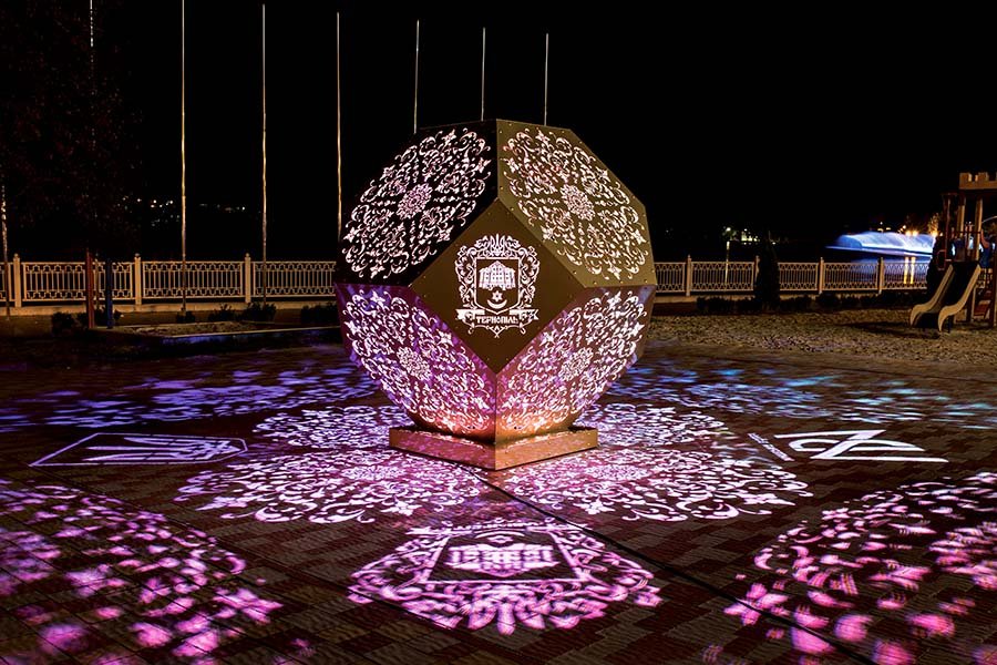 Decorative illuminated geometric sculpture projecting intricate floral patterns onto the ground in a public night installation Illuminated geometric sculpture with patterned light projections