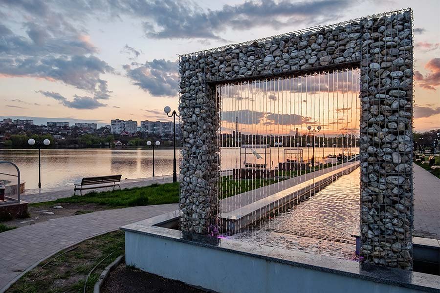 Stone-frame water curtain installation overlooking a riverside promenade at sunset